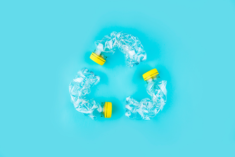 Three crushed plastic bottles with yellow caps arranged in a triangular shape to symbolize recycling, set against a bright blue background.